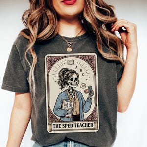 Puede incluir: Camiseta gris oscuro con un gr&aacute;fico estilo carta del tarot. El gr&aacute;fico representa a una profesora esqueleto sosteniendo una carpeta y un fidget spinner, con el texto "THE SPED TEACHER" en la parte inferior. El dise&ntilde;o est&aacute; enmarcado en un borde decorativo.