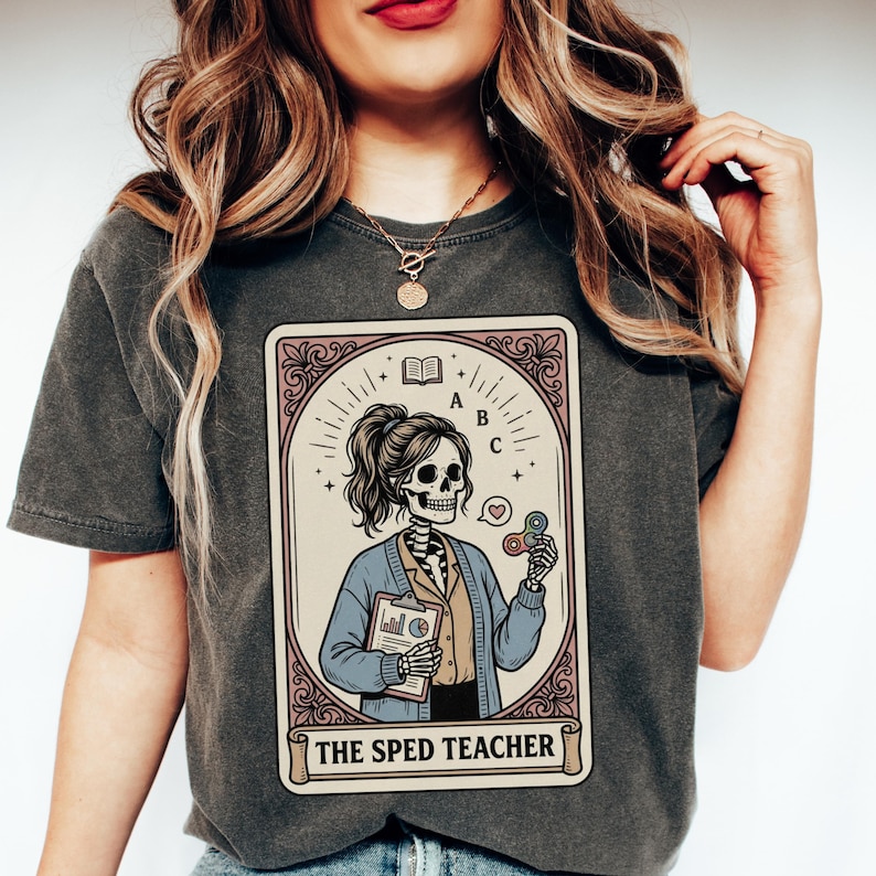 Puede incluir: Camiseta gris oscuro con un gr&aacute;fico estilo carta del tarot. El gr&aacute;fico representa a una profesora esqueleto sosteniendo una carpeta y un fidget spinner, con el texto "THE SPED TEACHER" en la parte inferior. El dise&ntilde;o est&aacute; enmarcado en un borde decorativo.