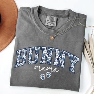 Puede incluir: Una camiseta de manga corta gris carb&oacute;n con la palabra BUNNY en letras grandes de bloque rellenas con un patr&oacute;n floral azul. Debajo de la palabra, el texto mama est&aacute; impreso en letra cursiva blanca, seguido de dos iconos de huellas de conejo.