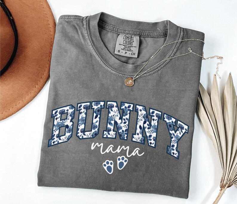 Puede incluir: Una camiseta de manga corta gris carb&oacute;n con la palabra BUNNY en letras grandes de bloque rellenas con un patr&oacute;n floral azul. Debajo de la palabra, el texto mama est&aacute; impreso en letra cursiva blanca, seguido de dos iconos de huellas de conejo.