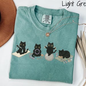 Puede incluir: Una camiseta de color verde claro con un gr&aacute;fico de cuatro gatos negros realizando diversos ejercicios, como yoga y pilates. La camiseta tiene cuello redondo y una etiqueta Comfort Colors. El texto "Light Green" aparece en la esquina superior derecha.