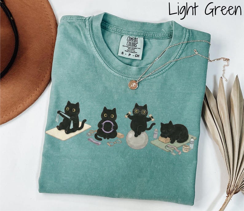 Puede incluir: Una camiseta de color verde claro con un gr&aacute;fico de cuatro gatos negros realizando diversos ejercicios, como yoga y pilates. La camiseta tiene cuello redondo y una etiqueta Comfort Colors. El texto "Light Green" aparece en la esquina superior derecha.