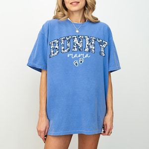Puede incluir: Una camiseta azul de manga corta con la palabra BUNNY en un estampado floral azul oscuro, la palabra mama en letra cursiva blanca y dos huellas de pata blancas centradas en el pecho. La prenda tiene cuello redondo y corte holgado.