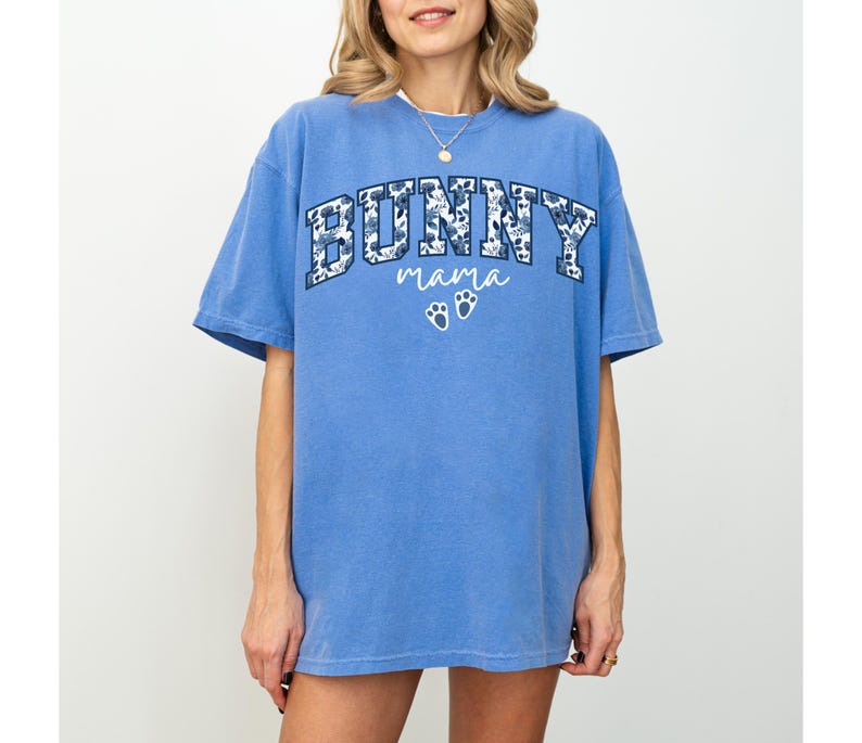 Puede incluir: Una camiseta azul de manga corta con la palabra BUNNY en un estampado floral azul oscuro, la palabra mama en letra cursiva blanca y dos huellas de pata blancas centradas en el pecho. La prenda tiene cuello redondo y corte holgado.