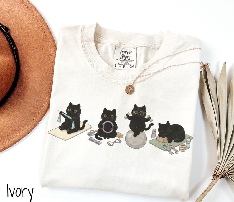 Puede incluir: Una camiseta color marfil con un gr&aacute;fico de cuatro gatos negros haciendo ejercicio. Un gato usa una banda de resistencia, otro sostiene un aro de pilates, otro se equilibra en una pelota de yoga con mancuernas y otro descansa en una esterilla. Etiqueta Comfort Colors.
