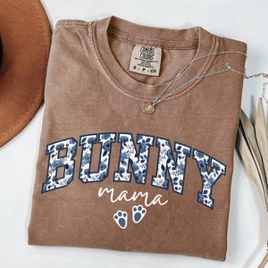 Puede incluir: Una camiseta marr&oacute;n de manga corta con cuello redondo. El frente presenta el texto BUNNY en letras grandes de bloque rellenas con un patr&oacute;n floral azul, seguido de la palabra mama y dos peque&ntilde;as huellas de pata blancas en letra cursiva.