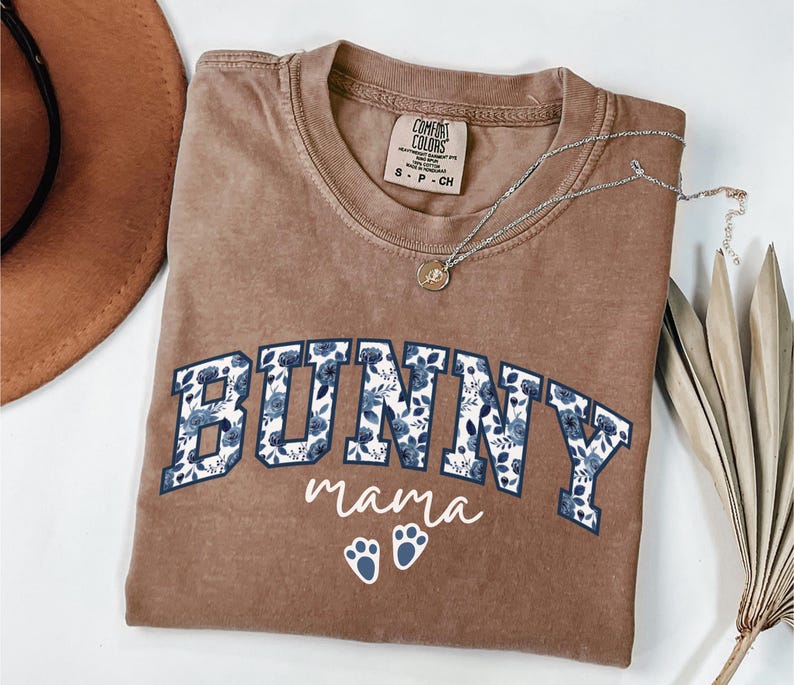 Puede incluir: Una camiseta marr&oacute;n de manga corta con cuello redondo. El frente presenta el texto BUNNY en letras grandes de bloque rellenas con un patr&oacute;n floral azul, seguido de la palabra mama y dos peque&ntilde;as huellas de pata blancas en letra cursiva.
