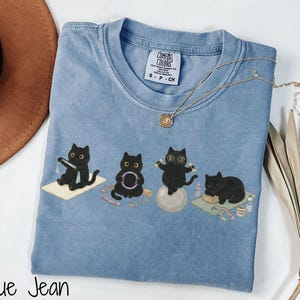 Puede incluir: Una camiseta color azul jean con un estampado gr&aacute;fico de cuatro gatos negros realizando varios ejercicios, incluyendo yoga y pilates. La camiseta presenta una etiqueta de la marca Comfort Colors en el cuello interior y cuello redondo.