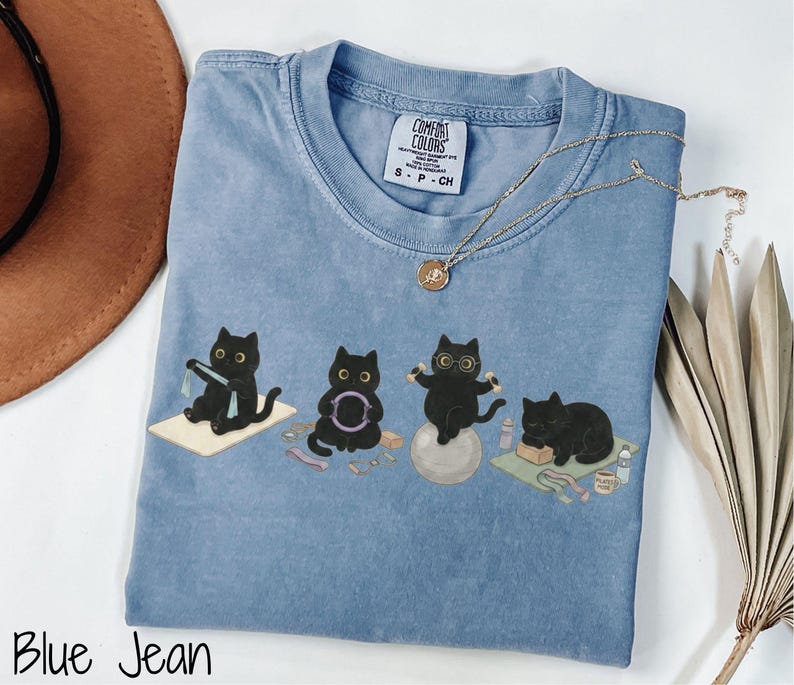 Puede incluir: Una camiseta color azul jean con un estampado gr&aacute;fico de cuatro gatos negros realizando varios ejercicios, incluyendo yoga y pilates. La camiseta presenta una etiqueta de la marca Comfort Colors en el cuello interior y cuello redondo.