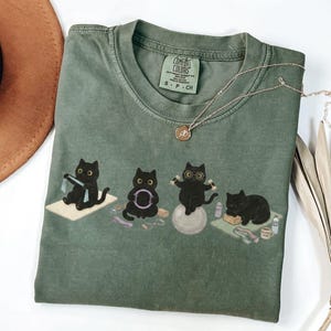 Puede incluir: Una camiseta verde oliva con un gr&aacute;fico de cuatro gatos negros realizando diversos ejercicios, incluyendo el uso de una banda de resistencia, un aro de pilates, una pelota de estabilidad con gafas y descansando sobre una esterilla de yoga.