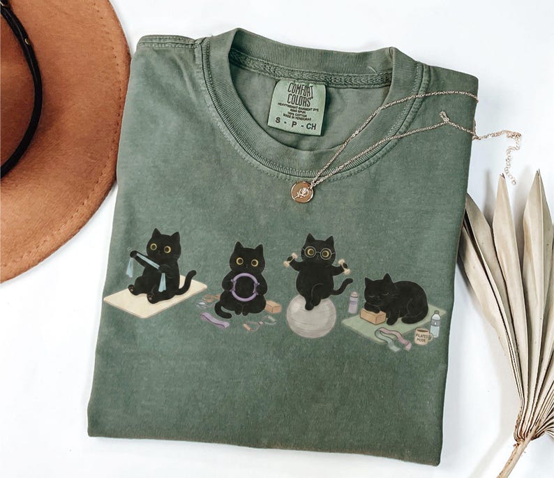 Puede incluir: Una camiseta verde oliva con un gr&aacute;fico de cuatro gatos negros realizando diversos ejercicios, incluyendo el uso de una banda de resistencia, un aro de pilates, una pelota de estabilidad con gafas y descansando sobre una esterilla de yoga.