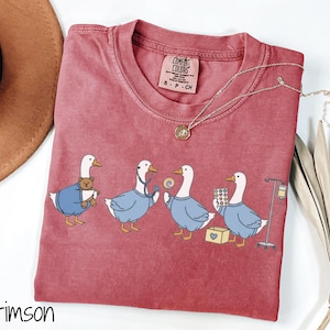 May include: Crimson Comfort Colors t-shirt featuring a whimsical illustration of four geese dressed as medical professionals. The geese are depicted with a teddy bear, stethoscope, lollipop, pills, and IV drip. A gold necklace is draped on the shirt.