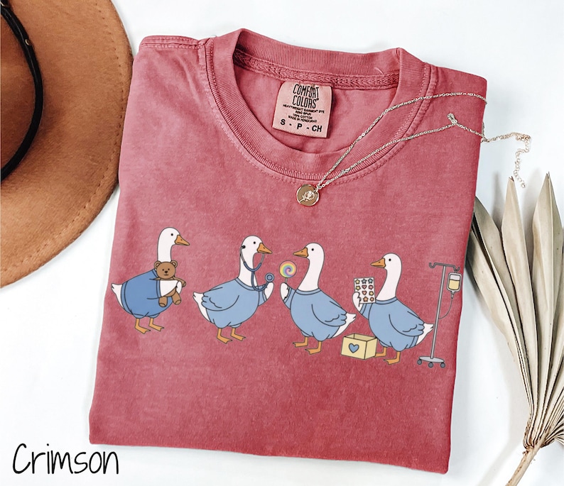 May include: Crimson Comfort Colors t-shirt featuring a whimsical illustration of four geese dressed as medical professionals. The geese are depicted with a teddy bear, stethoscope, lollipop, pills, and IV drip. A gold necklace is draped on the shirt.