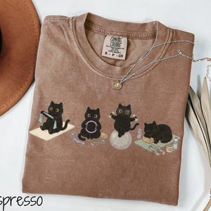 Puede incluir: Una camiseta color espresso con un estampado gr&aacute;fico de cuatro gatos negros realizando varios ejercicios de yoga y fitness. La camiseta cuenta con una etiqueta de la marca Comfort Colors en el cuello y un escote redondo. Los gatos aparecen usando esterillas de yoga, un aro de pilates, una pelota de estabilidad y un bloque de yoga.