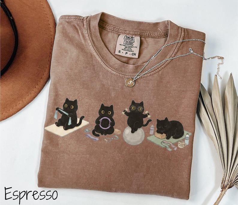 Puede incluir: Una camiseta color espresso con un estampado gr&aacute;fico de cuatro gatos negros realizando varios ejercicios de yoga y fitness. La camiseta cuenta con una etiqueta de la marca Comfort Colors en el cuello y un escote redondo. Los gatos aparecen usando esterillas de yoga, un aro de pilates, una pelota de estabilidad y un bloque de yoga.