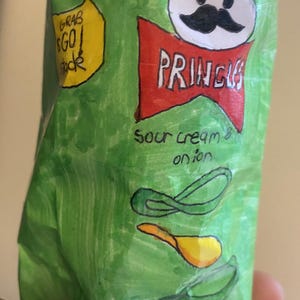 Könnte beinhalten: Eine grüne, zerknitterte Papiertüte mit einer Zeichnung eines lächelnden Comic-Gesichts mit einem Schnurrbart und einer roten Schleife. Die Tüte trägt die Aufschrift "PRINGLES" in roten Buchstaben und "Sour Cream & Onion" in grünen Buchstaben.