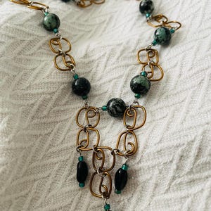May include: A gold and silver chain necklace with green and black beads. The necklace has a pendant with a green and black bead at the bottom.