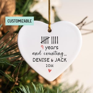 May include: White heart-shaped ceramic ornament with black text that reads "9 years and counting... DENISE & JACK 20XX" with a small pink heart at the bottom.