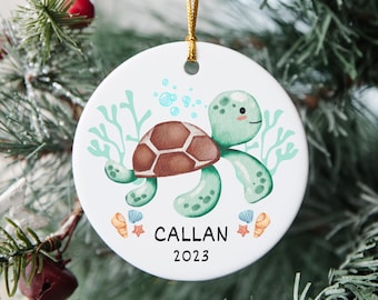 May include: A white round ceramic bauble featuring a green sea turtle illustration, surrounded by seaweed and bubbles. The bauble has the name "CALLAN" and the year "2023" printed on it.