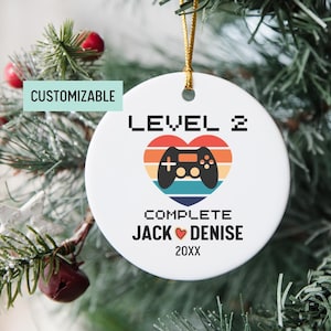May include: A white round ceramic ornament with a colorful retro-style video game controller design. The ornament says "Level 2 Complete Jack ♥ Denise 20XX".