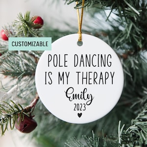 May include: White round ceramic Christmas ornament with black text that reads "Pole Dancing is My Therapy" and a personalized name "Emily" and the year "2023".