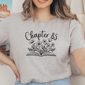 May include: Light gray t-shirt with the text "Chapter 85" above an open book with floral illustrations. The shirt is made of a soft material and is suitable for casual wear. The design is in black ink.