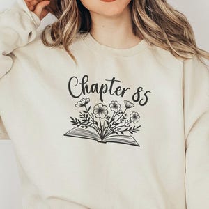 May include: Cream-colored sweatshirt with the text "Chapter 85" above a floral illustration. The design features an open book with flowers and leaves. The model is wearing a brown hat and blue jeans.