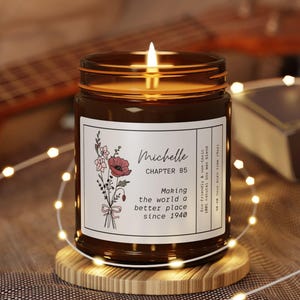 May include: A lit brown glass candle jar with a label that reads "Michelle Chapter 85 Making the world a better place since 1940." The label features floral illustrations. The candle sits on a wooden coaster, with fairy lights and a guitar in the background.