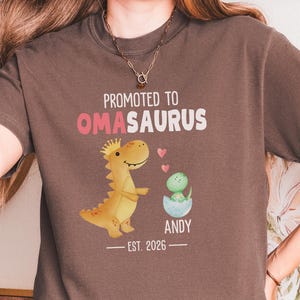 May include: A brown t-shirt with the text "Promoted to Omasaurus" in white and pink. A cartoon dinosaur and baby dinosaur are featured on the front. The text "Andy" and "Est. 2026" are also included.