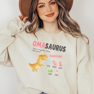 May include: A woman wearing a light beige sweatshirt with the text "OMASAURUS Like a regular Oma, but more RAWESOME!" and a graphic of a dinosaur with other smaller dinosaurs below.