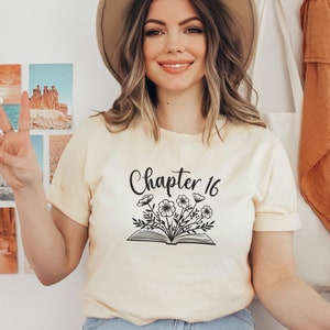 May include: A woman wearing a light yellow t-shirt with the text "Chapter 16" and a floral design above an open book. She is wearing a brown hat and blue jeans.