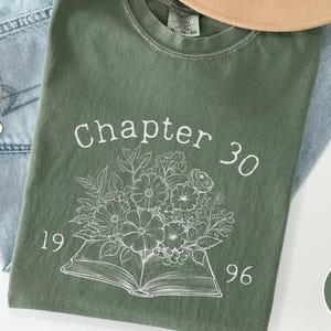 May include: A moss green t-shirt with the text "Chapter 30" and the years "1996" printed in white. A floral design is printed below the text. The shirt is laid on top of a pair of blue jeans and a tan hat.