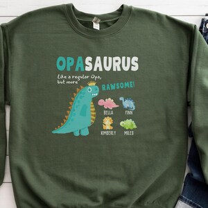 Peut inclure: Sweat-shirt vert avec un motif de dinosaure et le texte "OPASAURUS Like a regular Opa, but more RAWESOME!" avec les noms Bella, Finn, Kimberly et Miles sous le dinosaure.