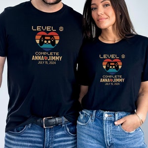 May include: Two black t-shirts with a retro video game controller graphic and the text "Level 8 Complete Anna Jimmy July 15, 2024".