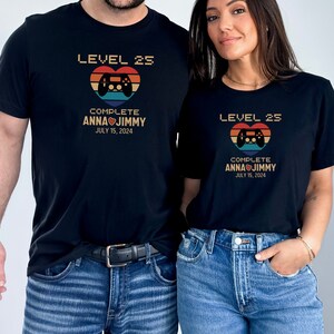 May include: A couple's matching black t-shirts with a retro video game controller graphic and the text "Level 25 Complete Anna Jimmy July 15, 2024".