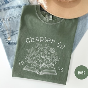Puede incluir: Una camiseta verde musgo con el texto "Chapter 50" y el año "1976" impresos en blanco. La camiseta presenta un diseño floral sobre un libro abierto. La camiseta está colocada sobre unos vaqueros azules y un sombrero beige.