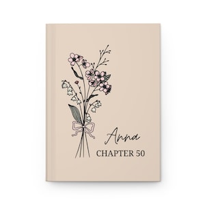 May include: A beige journal with a floral illustration of pink flowers and green leaves. The text "Anna Chapter 50" is printed on the front.