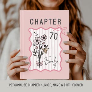 May include: A pink book cover with the word "CHAPTER" in dark grey at the top. The number "70" is centered, with the name "Emily" below it. A floral design with a bow is on the cover. The bottom of the image reads "PERSONALIZE CHAPTER NUMBER, NAME & BIRTH FLOWER."