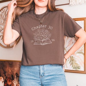 May include: A brown t-shirt with the text "Chapter 30" above a floral design and an open book. The numbers "19" and "96" are below the text. The shirt is paired with blue jeans.