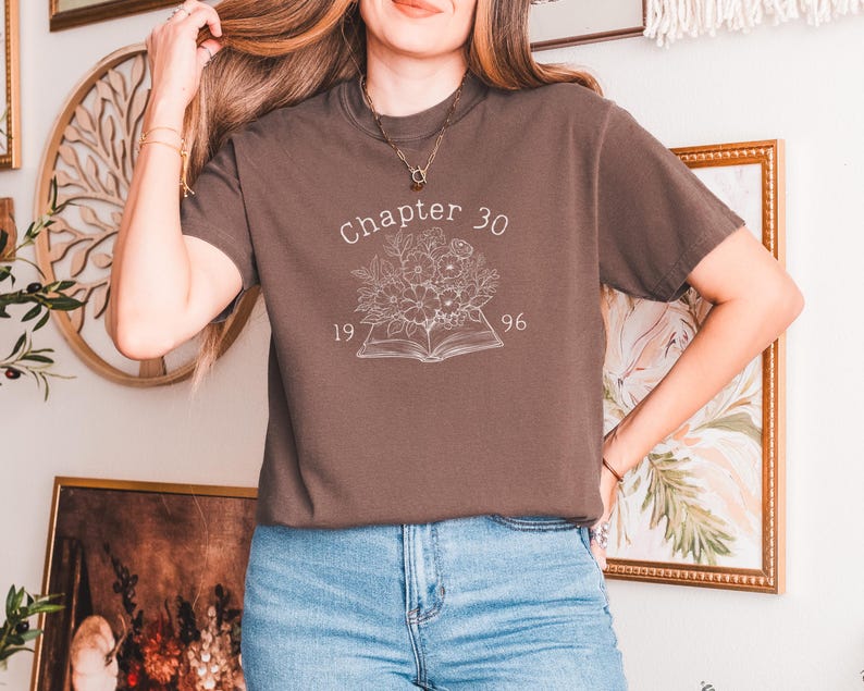 May include: A brown t-shirt with the text "Chapter 30" above a floral design and an open book. The numbers "19" and "96" are below the text. The shirt is paired with blue jeans.