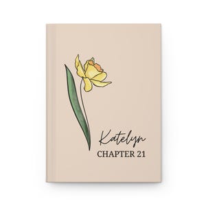 May include: A hardcover journal with a beige cover featuring a yellow daffodil illustration with a green stem and leaf. The name "Katelyn" is written in cursive, with "CHAPTER 21" below in a serif font.