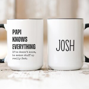 May include: Two white ceramic mugs with black handles and black rims. One mug says "Papi Knows Everything" and the other mug says "Josh".