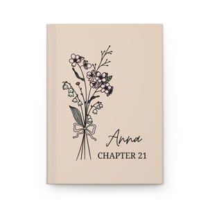 May include: A beige journal featuring a floral illustration of pink flowers and green leaves. The text "Anna Chapter 21" is printed on the front.