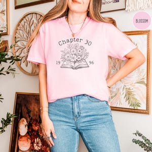 May include: A light pink t-shirt with the text "Chapter 30" above a floral design and the numbers "1996". The shirt is paired with blue jeans. The word "Blossom" is in a pink circle in the upper right corner.