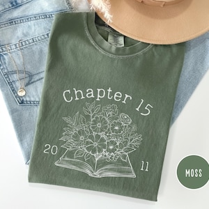 May include: A moss green t-shirt with the text "Chapter 15" and the numbers "2011" printed in white. A floral design is printed below the text. The shirt is laid on top of a pair of blue jeans and a tan hat.
