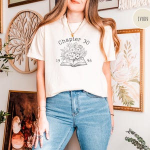 May include: Ivory-colored t-shirt with the text "Chapter 30" above a floral design and the numbers "1996". The shirt is paired with blue jeans. The shirt is a casual wear item.