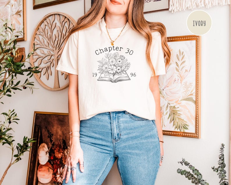 May include: Ivory-colored t-shirt with the text "Chapter 30" above a floral design and the numbers "1996". The shirt is paired with blue jeans. The shirt is a casual wear item.