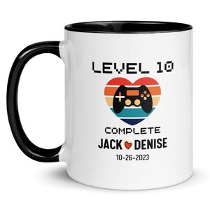 May include: White ceramic mug with a black rim and handle. The mug features a graphic of a video game controller in a heart with a retro sunset background. The text "LEVEL 10 COMPLETE JACK ♥ DENISE 10-26-2023" is printed on the mug.