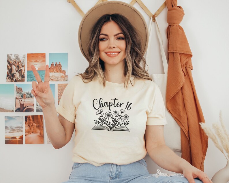 May include: A woman wearing a light yellow t-shirt with the text "Chapter 16" and a floral design above an open book. She is wearing a brown hat and blue jeans.