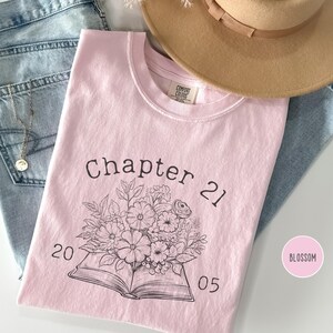 Może przedstawiać: Jasnoróżowa koszulka z napisem "Chapter 21" nad kwiatową ilustracją i liczbami "20 05". Koszulka leży na niebieskich dżinsach i beżowym kapeluszu.